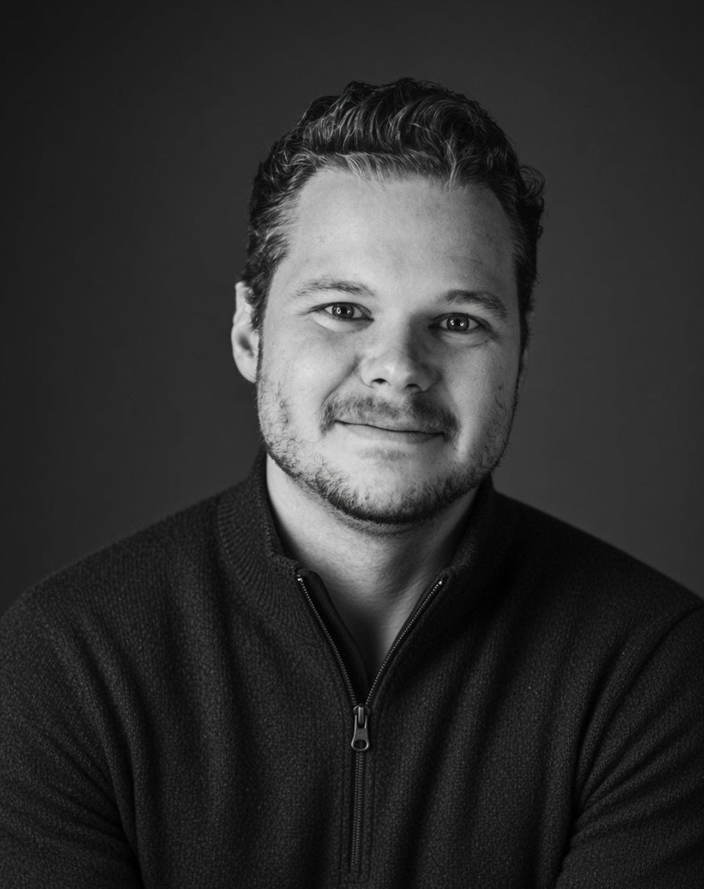 A black and white portrait of a man with short dark hair and a light beard, wearing a dark quarter-zip pullover sweater, with his arms crossed, set against a dark gray studio background.